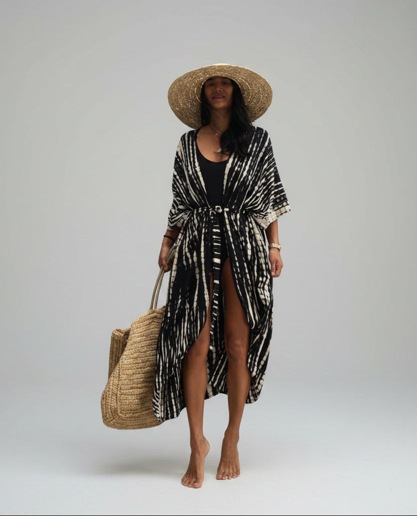 Woman in a black and white patterned dress with a straw hat and bag against a textured wall.