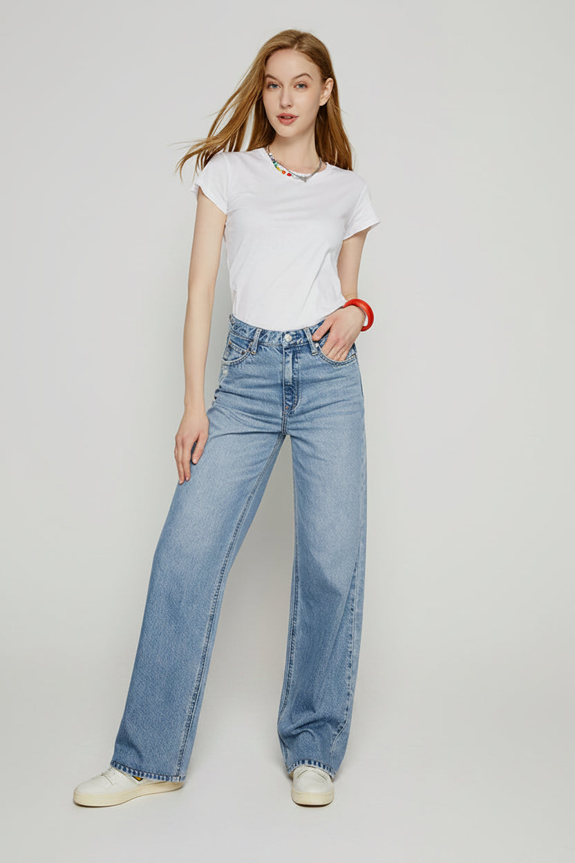 Woman wearing a white t-shirt and blue jeans standing on a patterned rug with a plain wall background