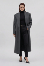 Person wearing a long dark coat and holding a brown leather bag, standing in front of a black vehicle.