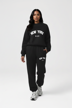 A model wearing a black sweatshirt and matching sweatpants set with white 'NEW YORK U.S.