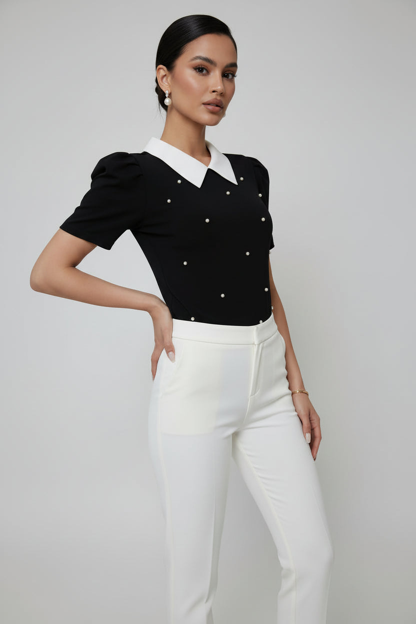 Woman wearing a black blouse with white collar and buttons, standing against a white curtain.