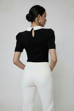 Woman wearing a black polka dot blouse with a white collar and white pants on a plain background