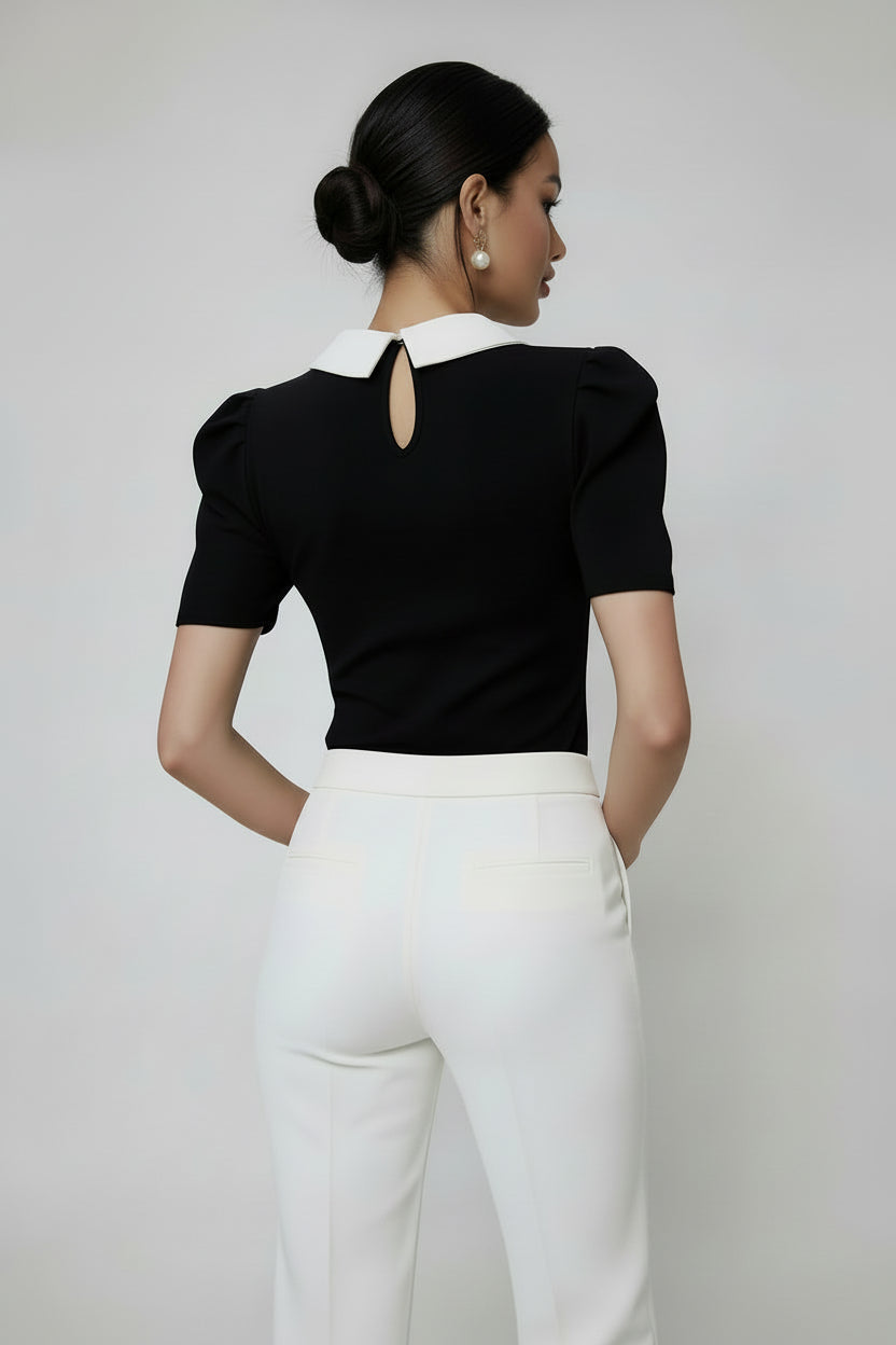 Woman wearing a black polka dot blouse with a white collar and white pants on a plain background