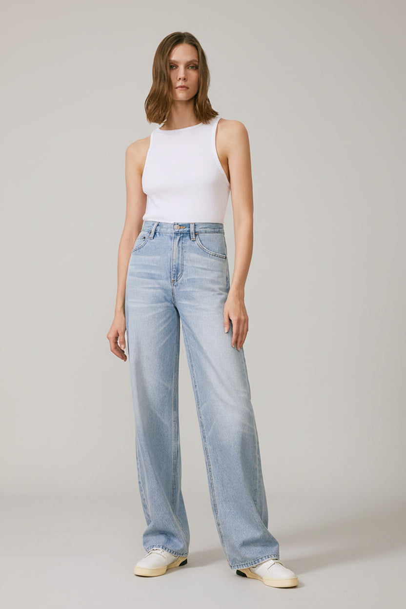 Woman wearing a white sleeveless top and light blue jeans standing on a patterned rug with a plain wall background.