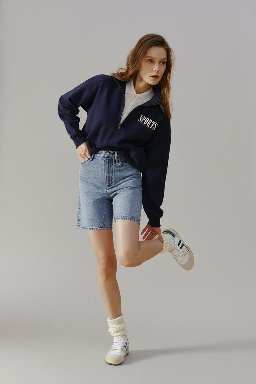 Woman wearing a navy blue sweatshirt with 'SPORTS' text, denim shorts, and white sneakers on a beige background