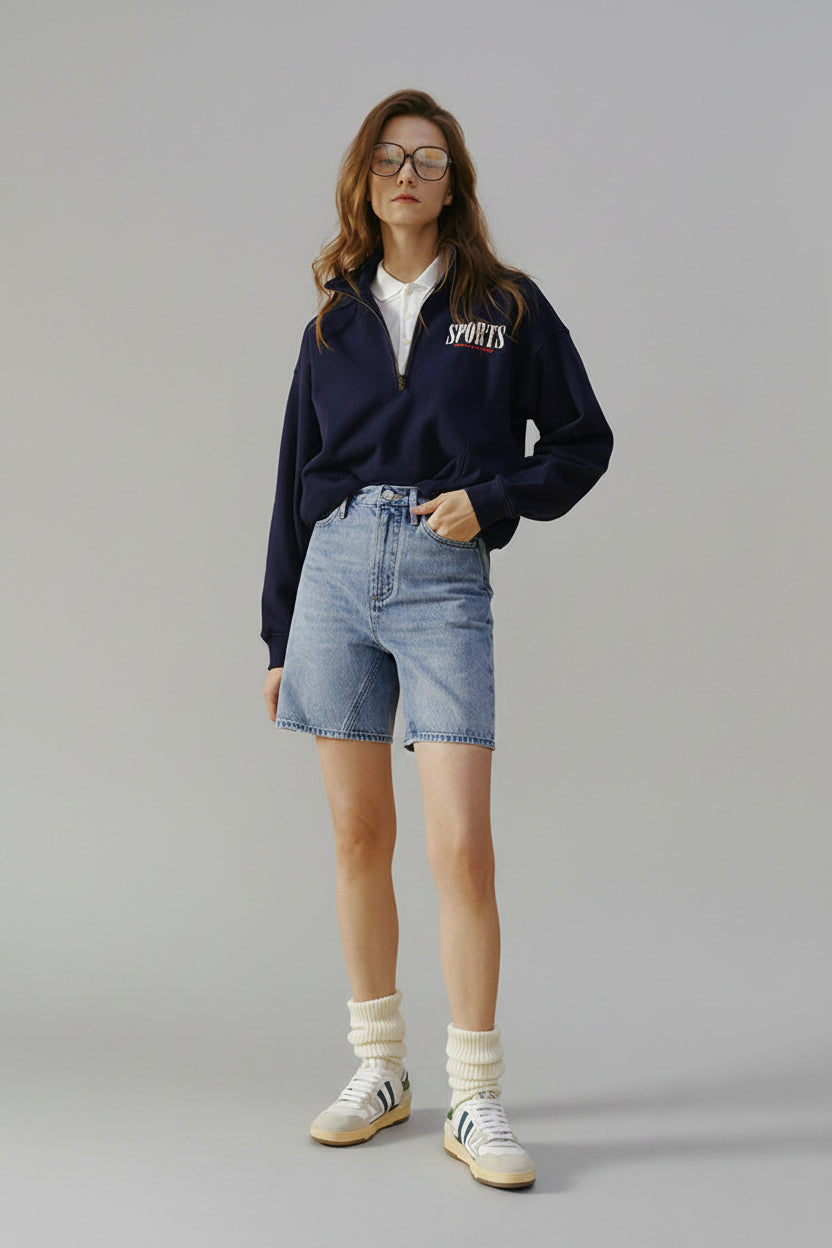 Woman wearing a navy sweatshirt with 'SPORTS' text, white shirt, blue shorts, and white sneakers on a beige background