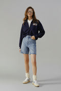 Woman wearing a navy sweatshirt with 'SPORTS' text, white shirt, blue shorts, and white sneakers on a beige background
