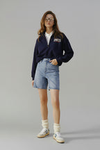 Woman wearing a navy sweatshirt with 'SPORTS' text, white shirt, blue shorts, and white sneakers on a beige background