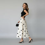 Woman wearing a black off-shoulder top and white polka dot skirt against a gray wall.