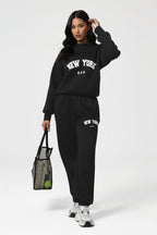 A model wearing a black sweatshirt and matching jogger pants set with white 'NEW YORK' graphics, holding a black mesh tote bag against a light gray studio background.