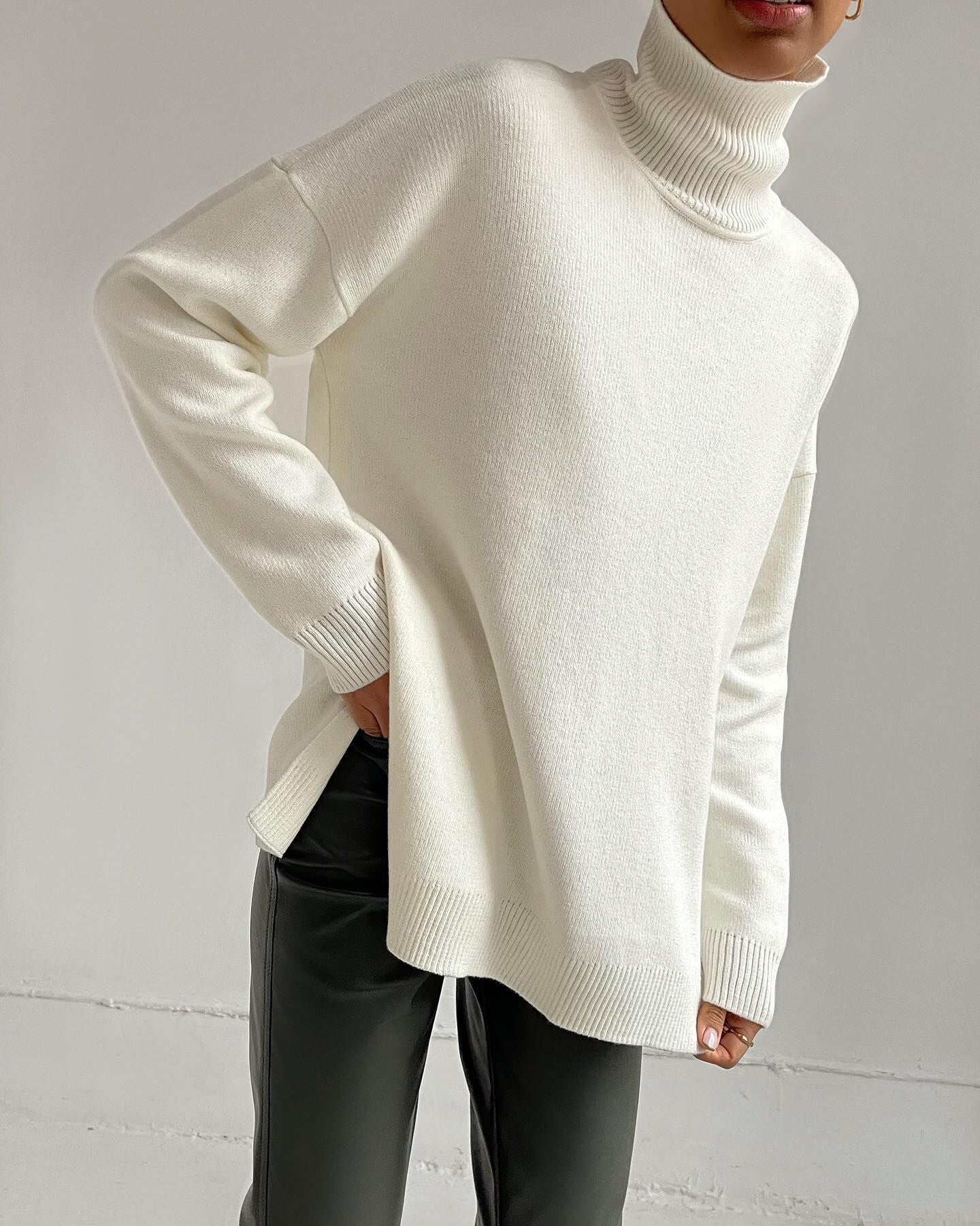 Person wearing a white turtleneck sweater and dark pants against a gray background