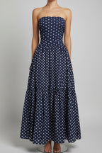 Woman wearing a strapless navy blue dress with white polka dots, holding a straw bag, standing against a white wall.
