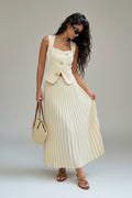 Woman in a white pleated skirt and vest standing against a stone wall.