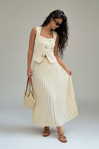 Woman in a white pleated skirt and vest standing against a stone wall.