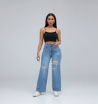 A woman standing in a room with white furniture, wearing a black top and blue distressed jeans with a high waist rise.