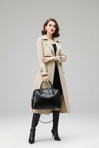 Black handbag held by a person wearing a beige coat and black boots on a blurred outdoor background.