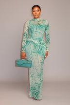 Woman in a green patterned outfit holding a matching handbag against a wooden wall.