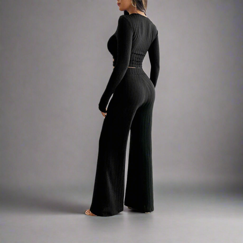 Woman wearing a black ribbed jumpsuit in a room with white walls and curtains.