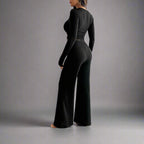 Woman wearing a black ribbed jumpsuit in a room with white walls and curtains.