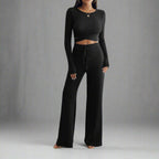 Person wearing a black long-sleeve top and wide-leg pants in a minimalistic room.