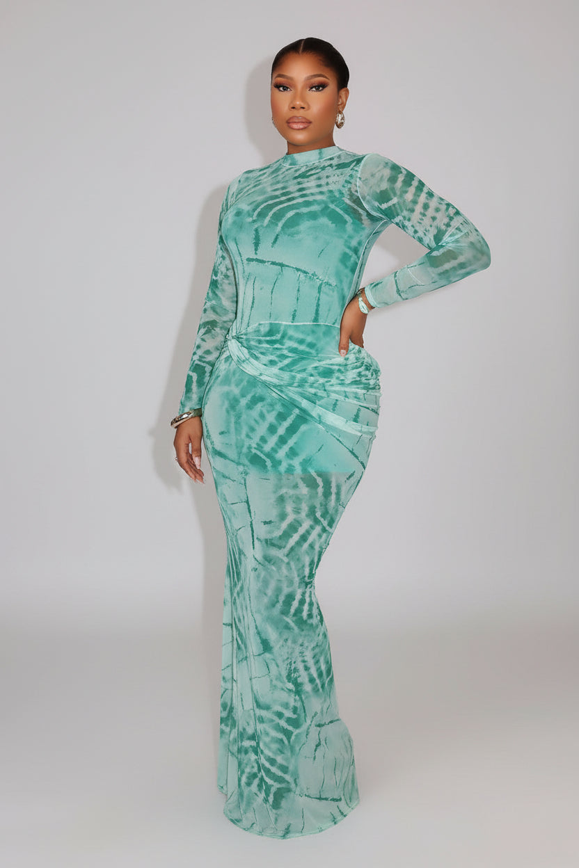 Woman in a teal patterned dress posing against a warm-lit wall.