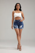 Woman wearing a white crop top and blue denim shorts in a minimalistic room with a chair and plant.
