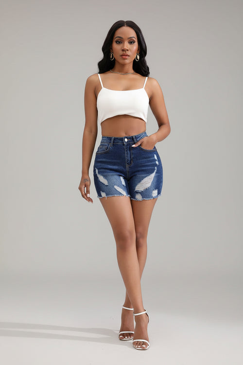 Woman wearing a white crop top and blue denim shorts in a minimalistic room with a chair and plant.