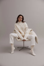 Woman wearing a white cable knit sweater sitting on a white chair.