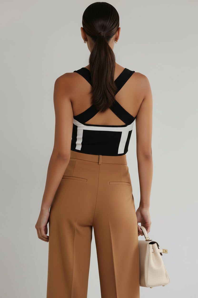 Woman wearing a black and white patterned top with tan pants, holding a cream handbag on a gray background