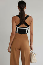 Woman wearing a black and white patterned top with tan pants, holding a cream handbag on a gray background