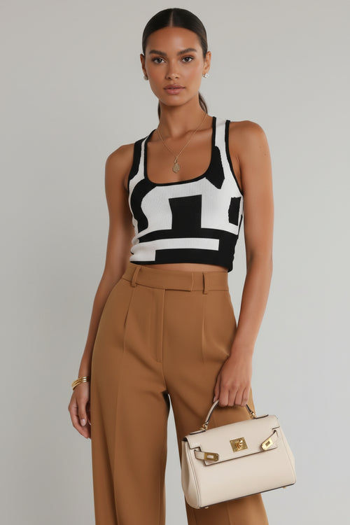 Woman wearing a black and white patterned top with tan pants, holding a beige handbag on a gray background