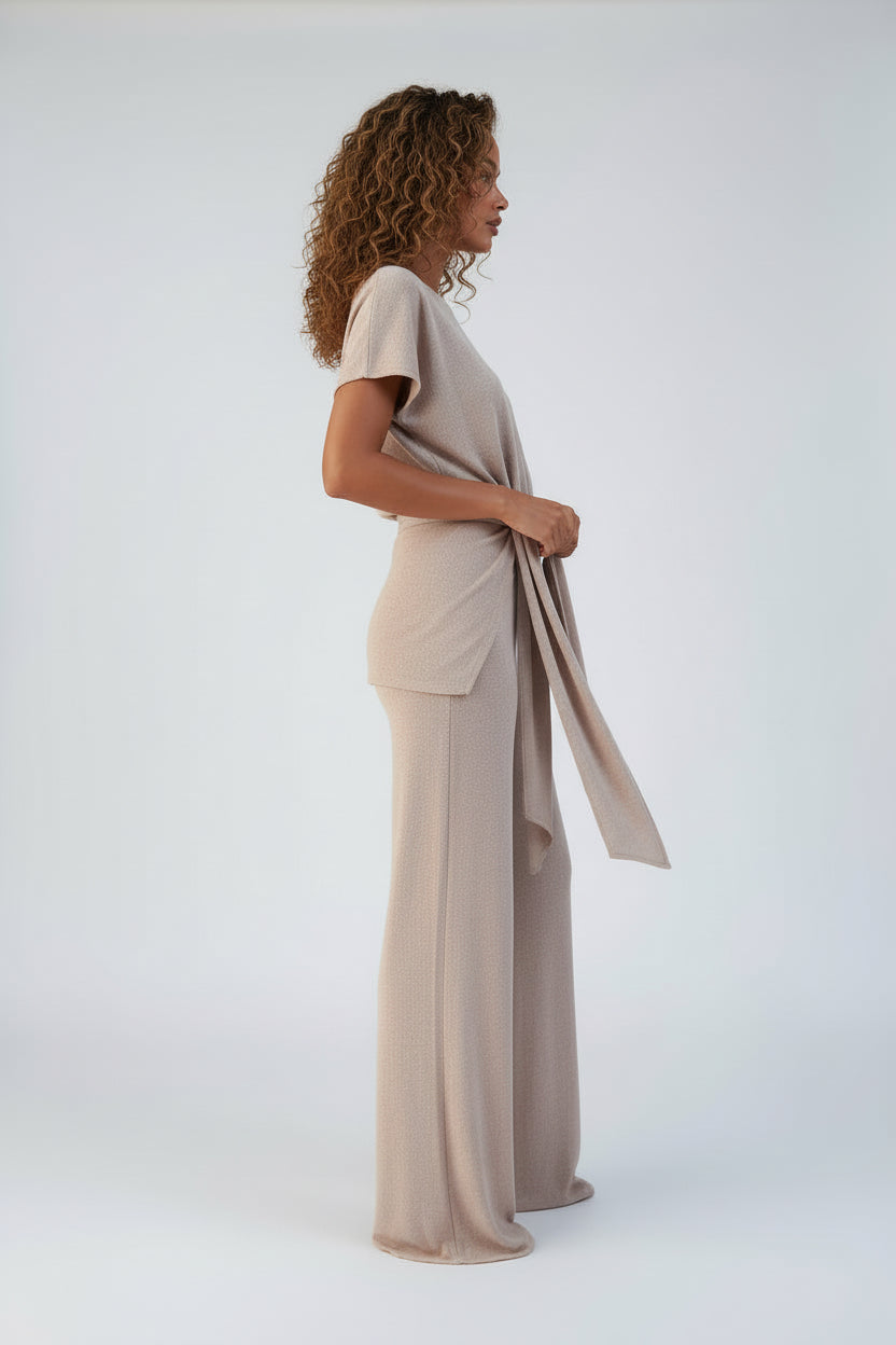 Person wearing a beige jumpsuit holding a phone and a handbag indoors.