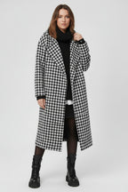 Person wearing a black and white houndstooth patterned coat against a beige wall.
