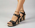 Black wedge sandals with cork soles worn by a person on a wooden floor.